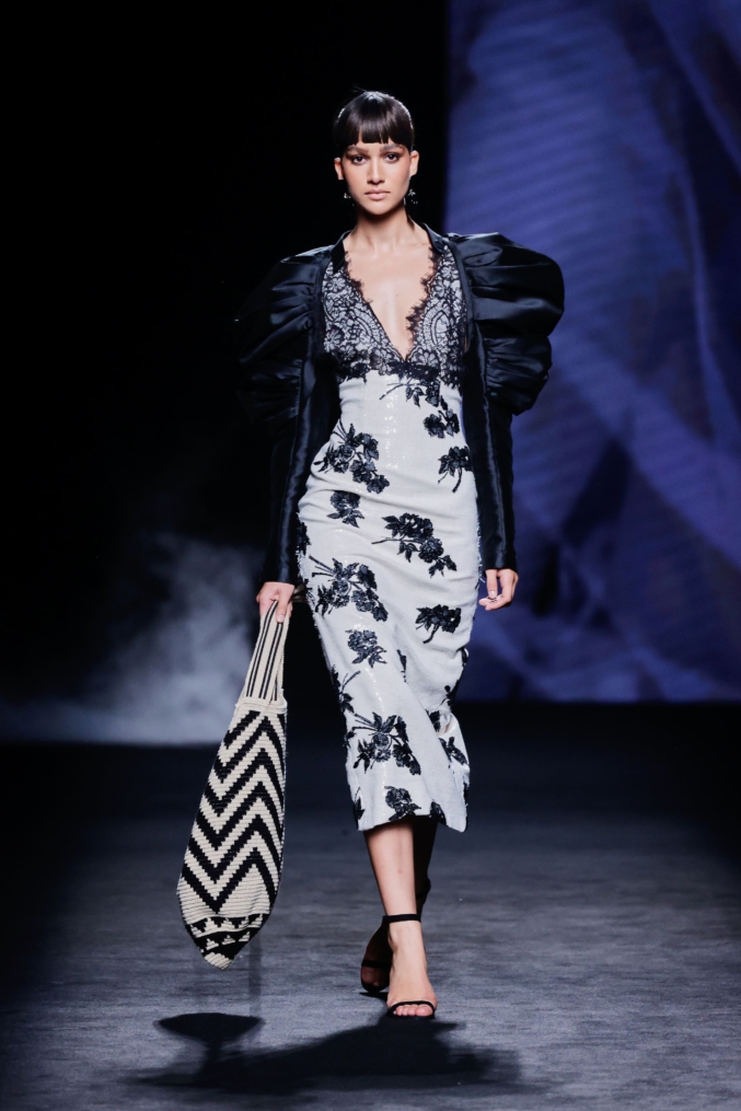 Model on the catwalk wearing a white midi dress by Malne decorated with black floral embroidery and a deep black lace neckline.