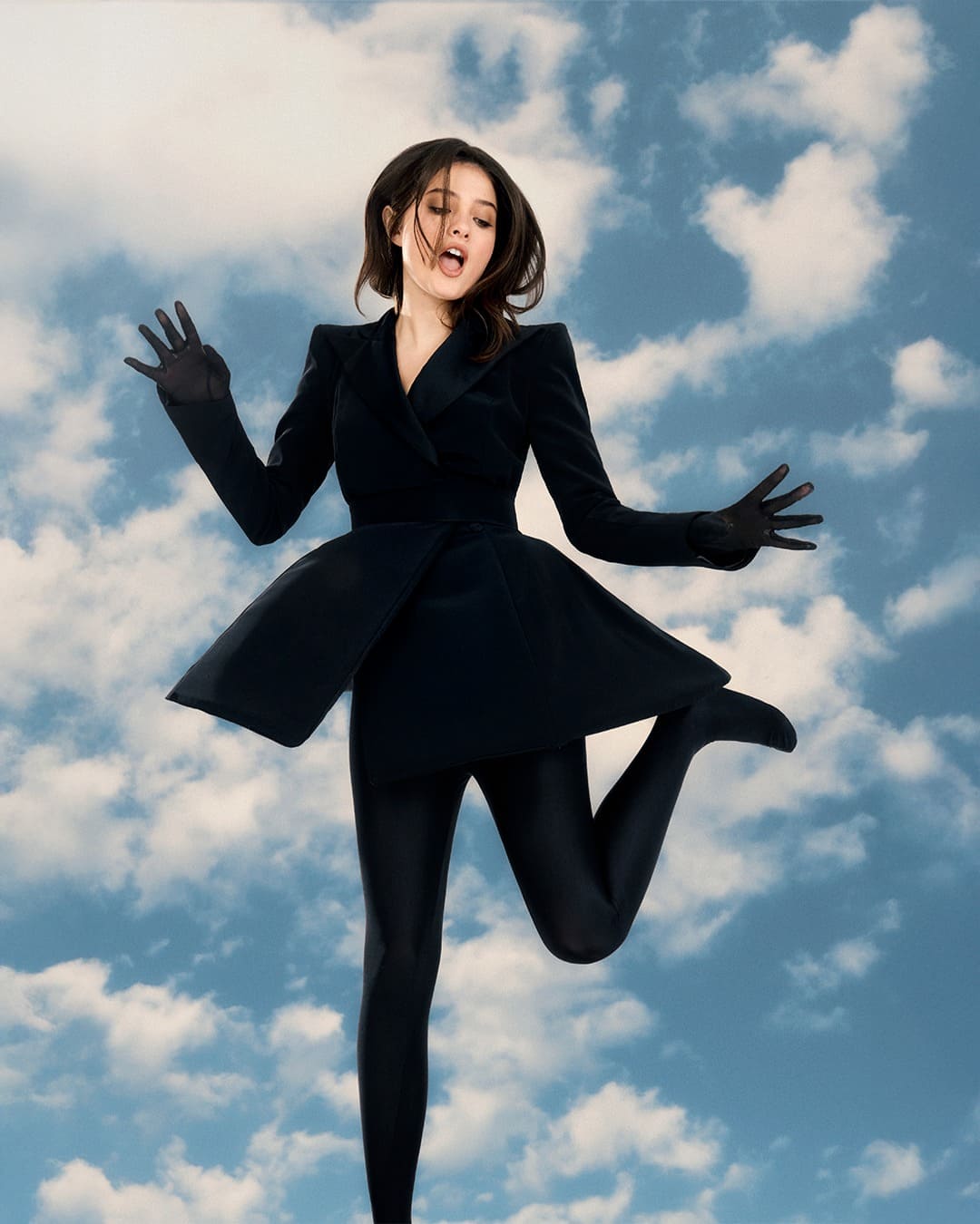 Mujer salta en el aire frente a un cielo azul con nubes blancas mientras lleva un conjunto negro estructurado con falda corta acampanada, guantes y medias ajustadas
