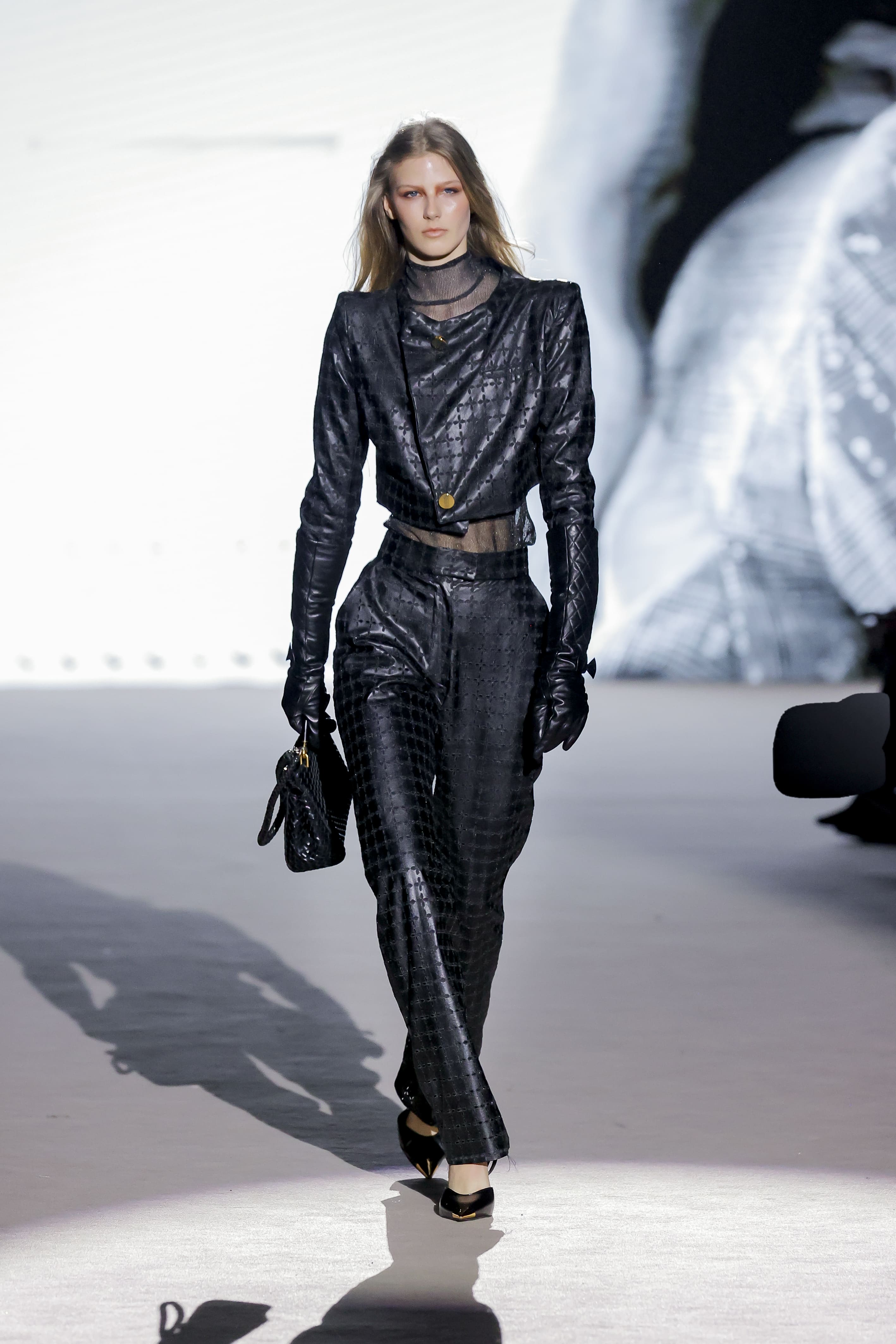 Modelo con conjunto negro ajustado con textura brillante de Malne en la Mercedes-Benz Fashion Week Madrid marzo de 2026 con guantes y bolso.