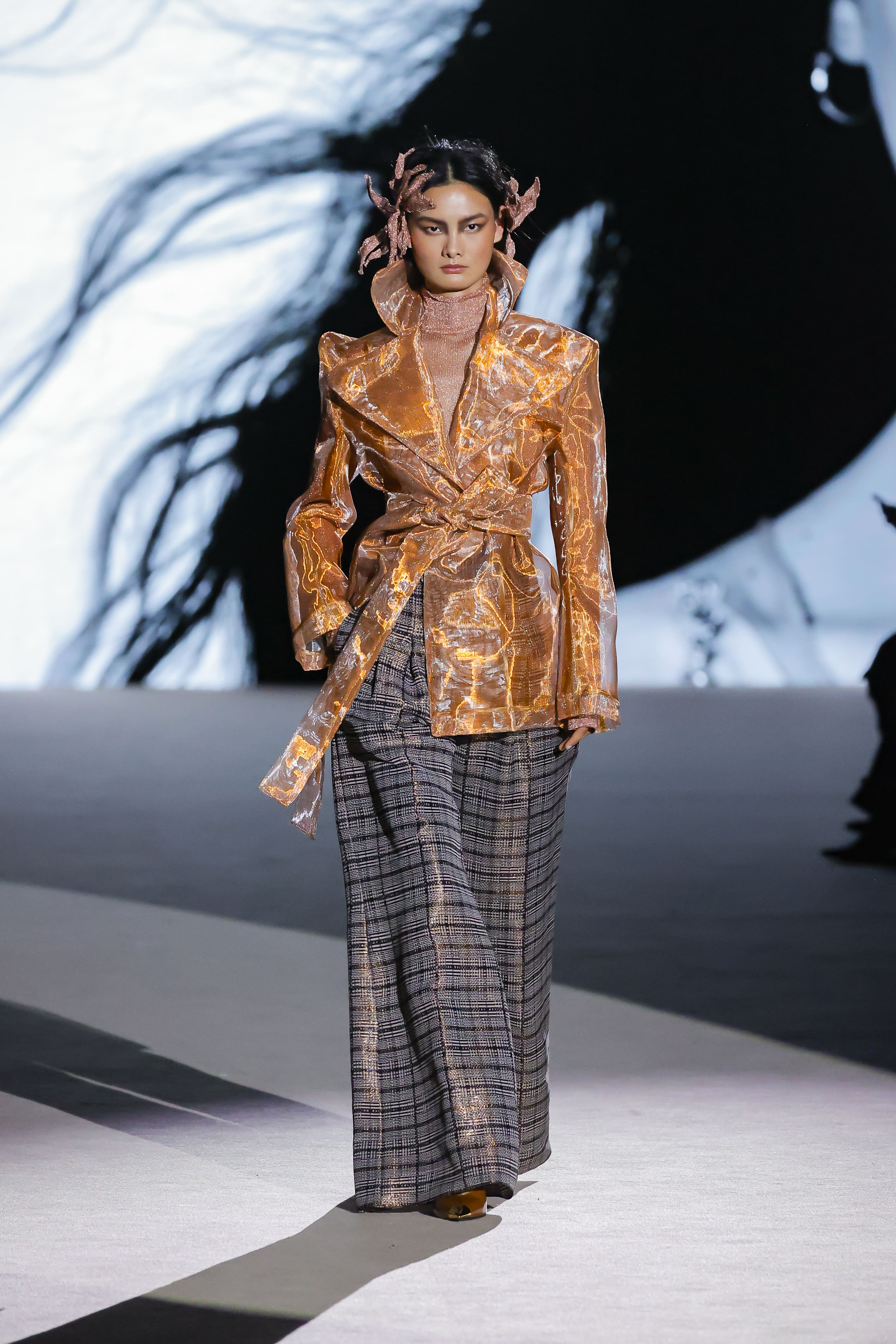Modelo con chaqueta brillante cobre y pantalón de cuadros de Malne en la Mercedes-Benz Fashion Week Madrid marzo de 2026 con cuello alto.