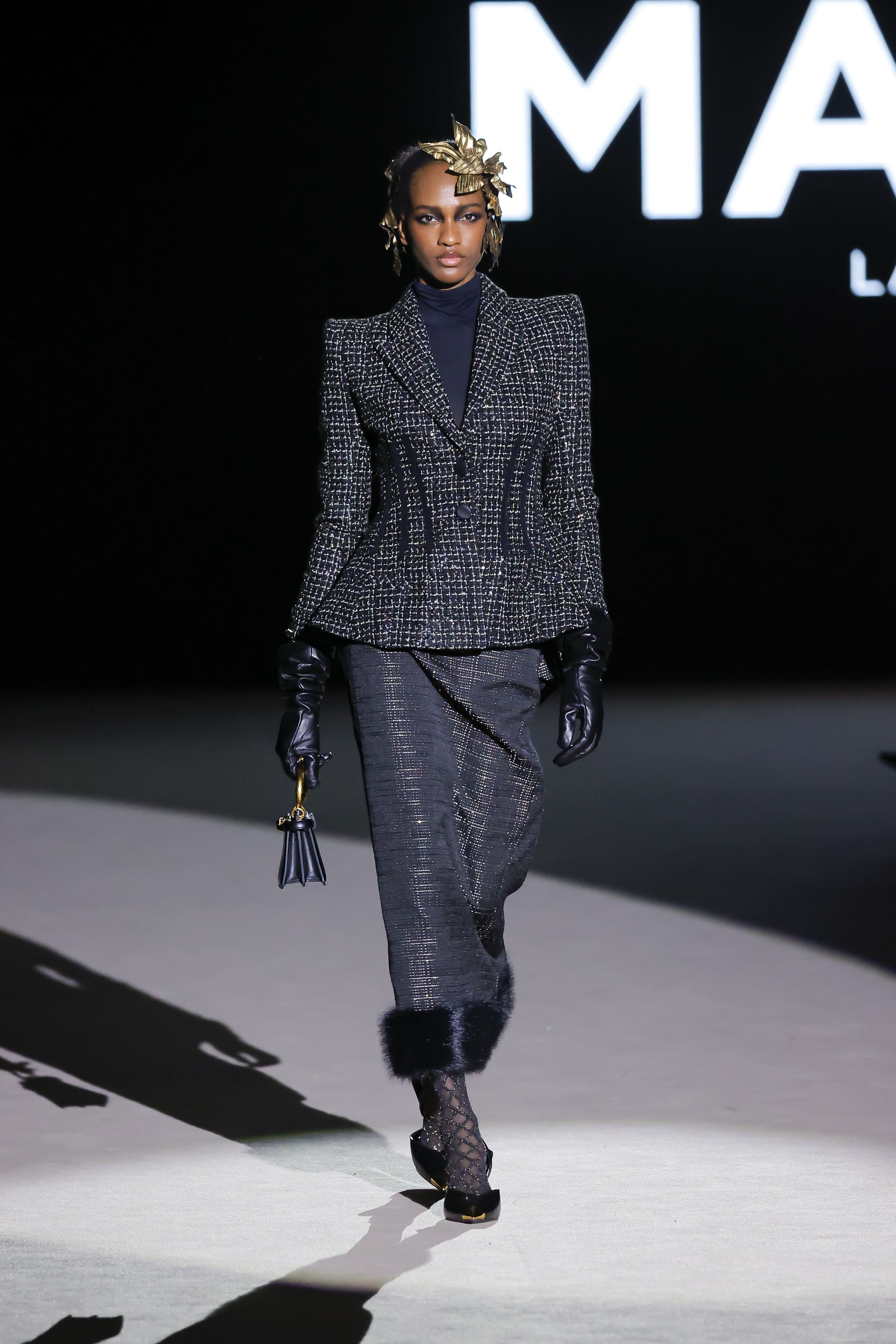 Modelo con traje estructurado gris de Malne en la Mercedes-Benz Fashion Week Madrid marzo de 2026 con hombreras marcadas guantes negros y tocado dorado.