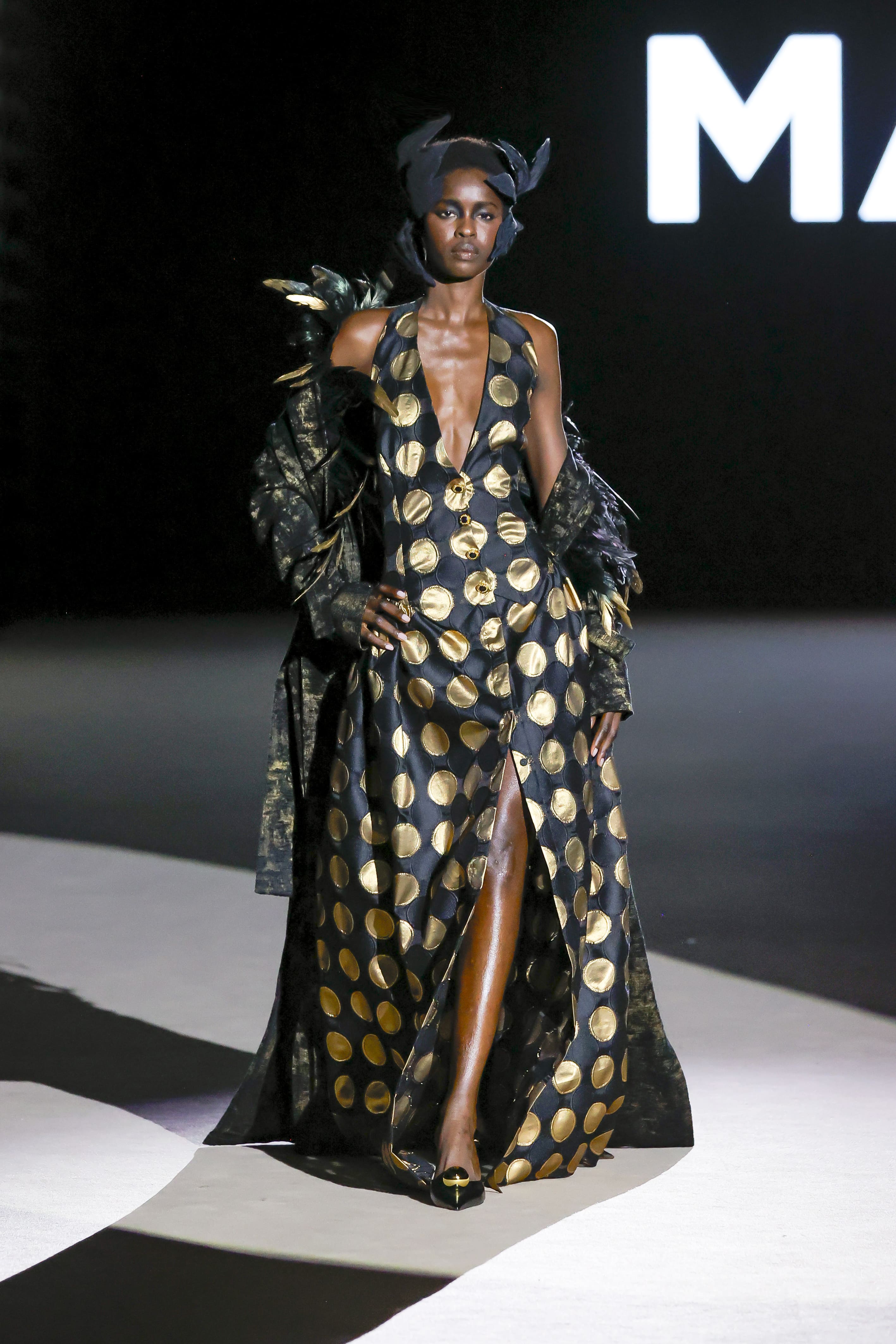 Modelo desfilando con diseño de Malne en la Mercedes-Benz Fashion Week Madrid en marzo de 2026 con vestido negro largo de lunares dorados y abrigo con plumas en los hombros.