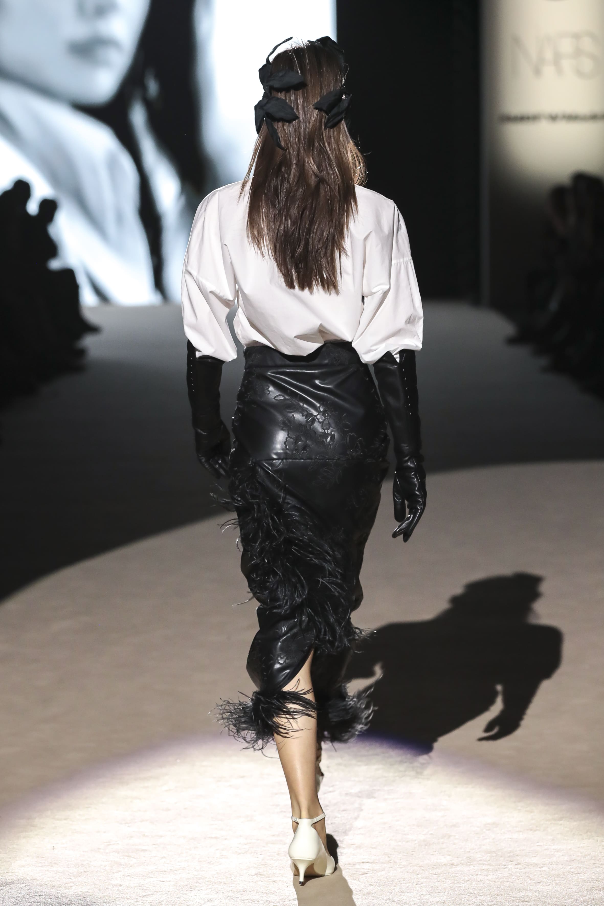 Modelo desfilando de espaldas con diseño de Malne en la Mercedes-Benz Fashion Week Madrid marzo de 2026 con camisa blanca falda negra brillante y guantes largos.