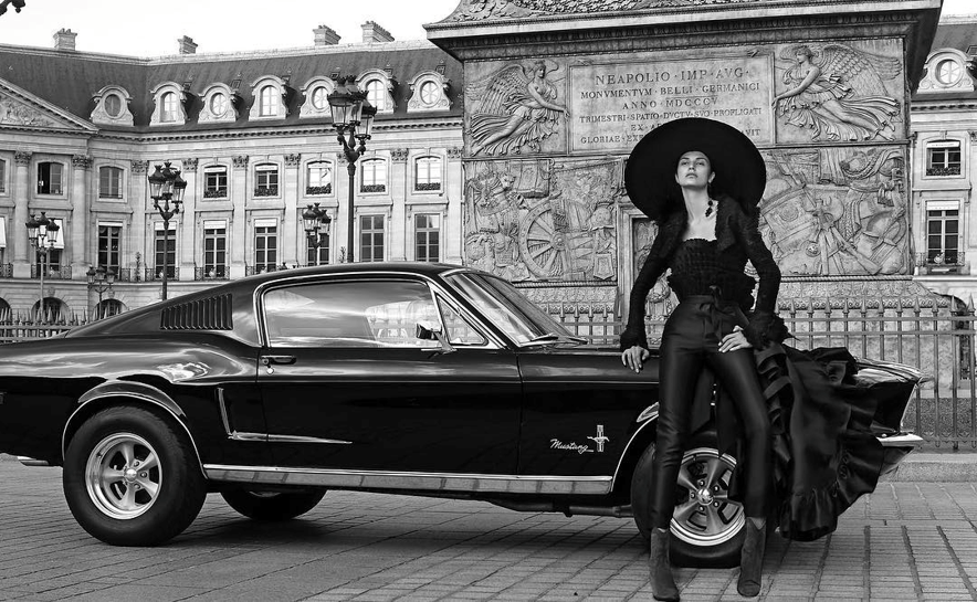 Modelo Malne vestida de negro junto a un coche clásico frente a un monumento urbano.