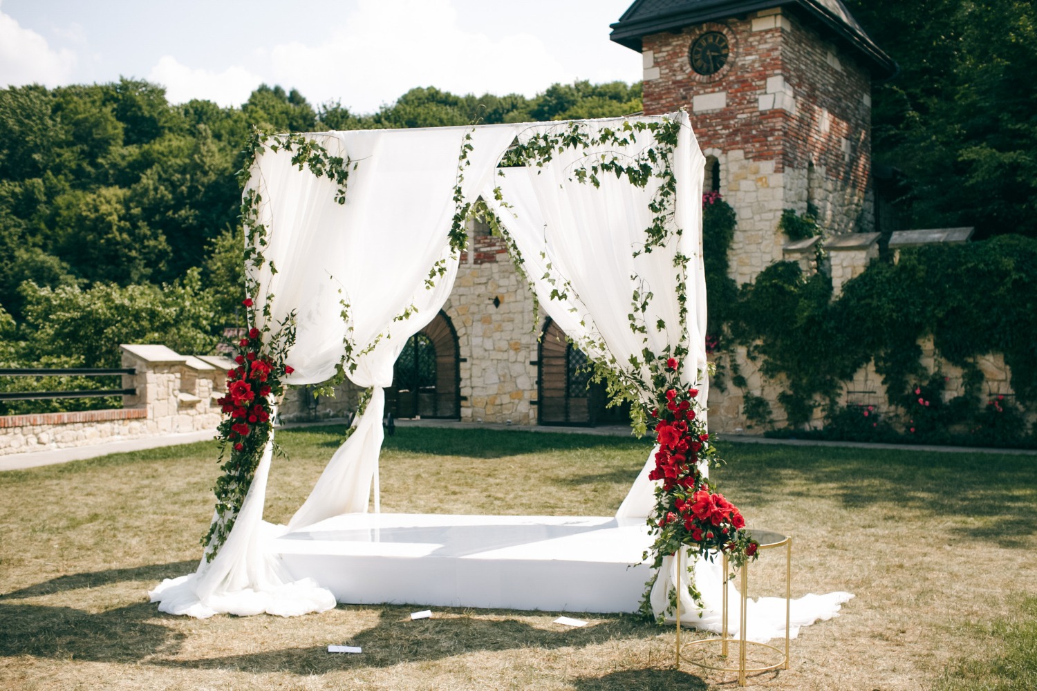 Dosel blanco para ceremonia de boda decorado con flores rojas y verdes frente a un edificio de piedra.